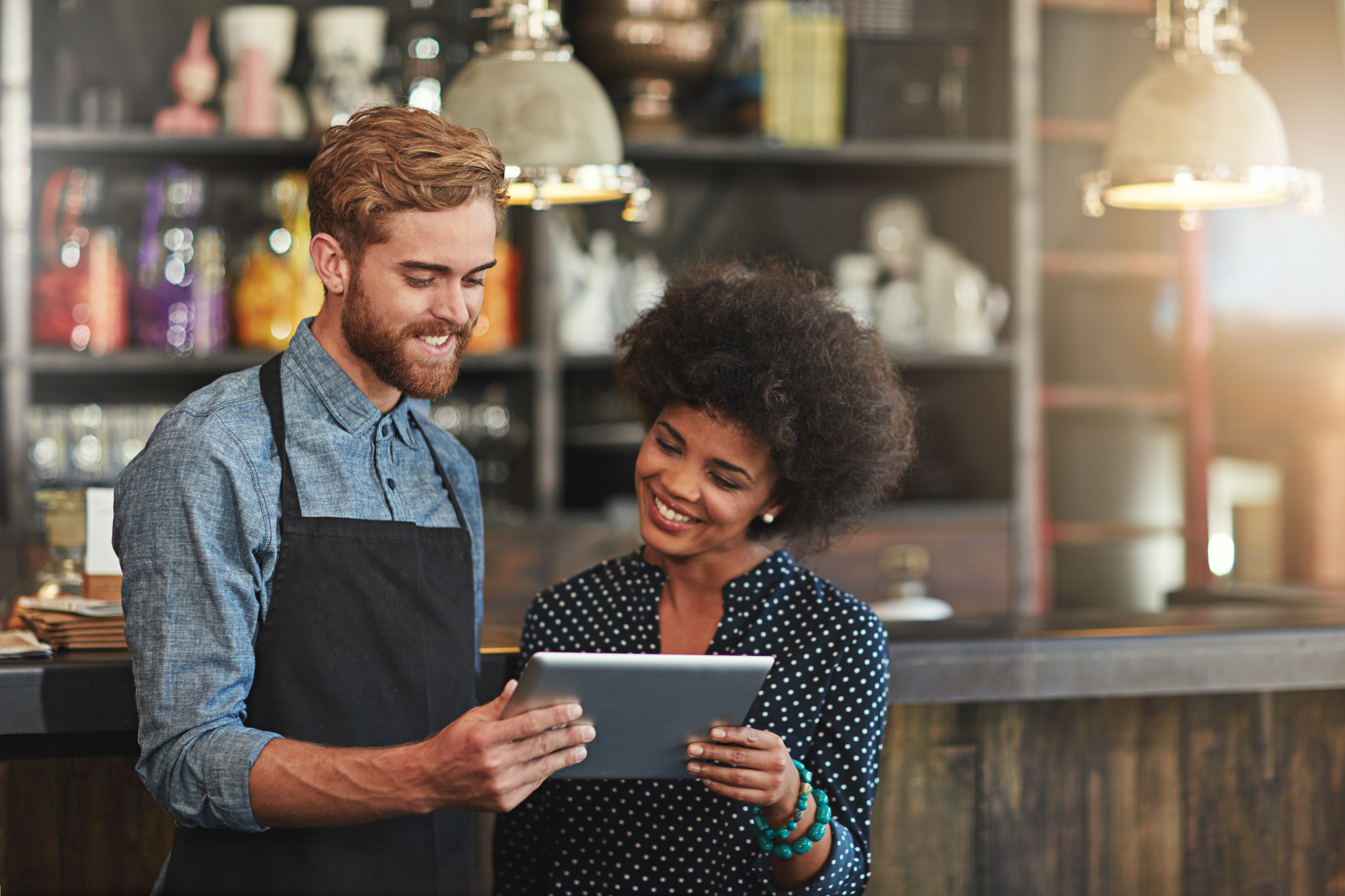 A cheerful bartender in a denim shirt and apron shares a tablet screen with a smiling sales woman with a curly afro.